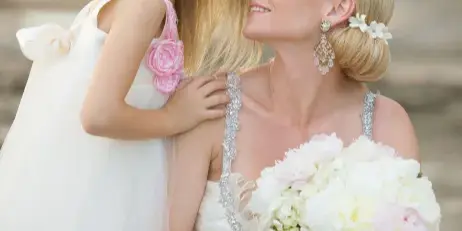 A bride with a styled blonde updo and floral hair accessory holding a white bouquet, embracing a young girl in a white dress.