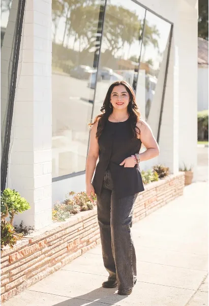 A woman with long dark hair wearing a black sleeveless top and jeans stands smiling in front of a modern building with large glass windows and a brick planter.