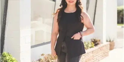 A woman with long dark hair wearing a black sleeveless top and jeans stands smiling in front of a modern building with large glass windows and a brick planter.