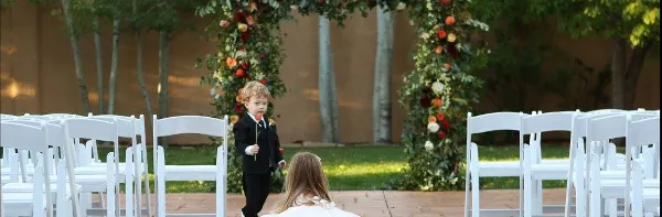 A young flower boy in a black suit walks down the aisle toward a bride during an outdoor wedding ceremony with white chairs and floral arches.