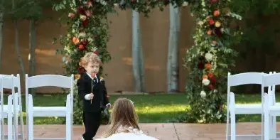 A young flower boy in a black suit walks down the aisle toward a bride during an outdoor wedding ceremony with white chairs and floral arches.