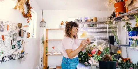 A florist arranging a bouquet in a bright studio workspace filled with fresh flowers and supplies.