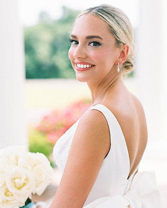 Bride in a white dress smiling toward the camera, with bridal makeup and a bouquet.