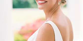 Bride in a white dress smiling toward the camera, with bridal makeup and a bouquet.