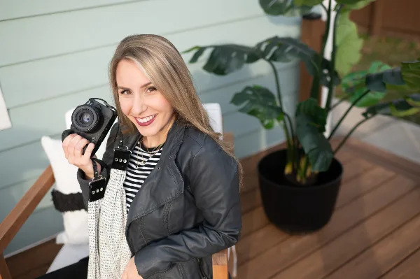 A smiling woman holding a professional camera, seated on a porch with a potted plant.