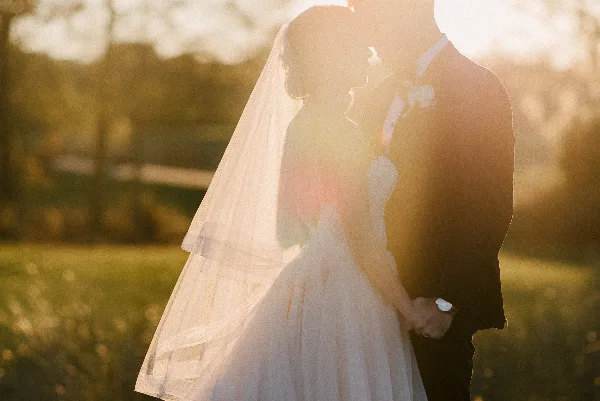 A bride and groom holding hands in a field during golden hour, with the bride's veil illuminated by the sun.