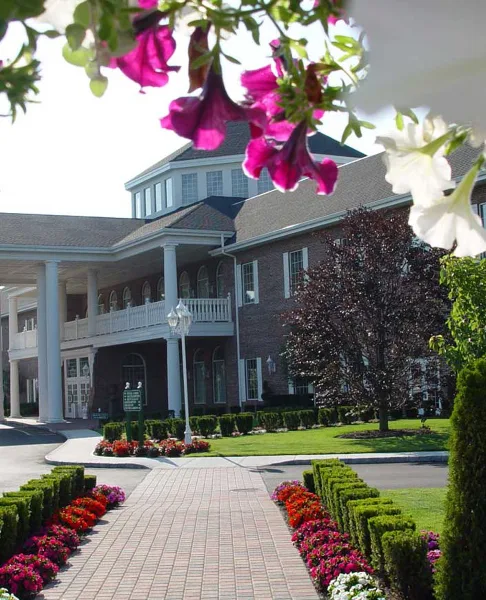 Exterior view of a large brick hotel building with a white columned porch, surrounded by a brick walkway, manicured hedges, and flower beds.