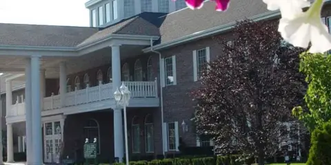 Exterior view of a large brick hotel building with a white columned porch, surrounded by a brick walkway, manicured hedges, and flower beds.