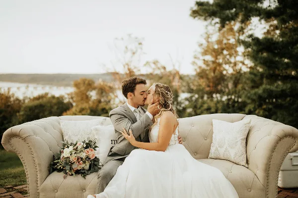 Bride and groom kissing on a tufted outdoor sofa with a bouquet and scenic backdrop.