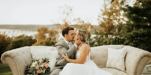 Bride and groom kissing on a tufted outdoor sofa with a bouquet and scenic backdrop.