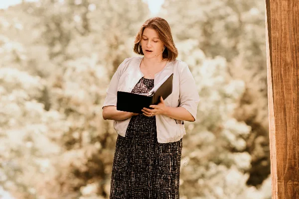 A woman standing outdoors reading from a book, likely officiating a ceremony.