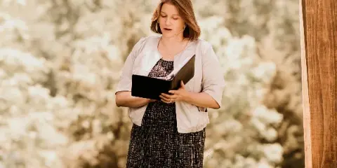 A woman standing outdoors reading from a book, likely officiating a ceremony.