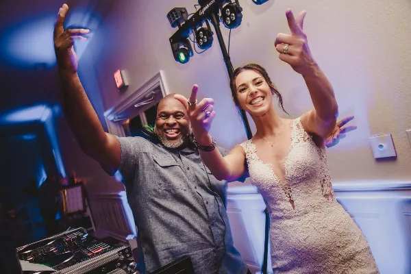 A smiling DJ and a bride in a white lace dress posing with raised hands in a venue with stage lighting and DJ equipment.