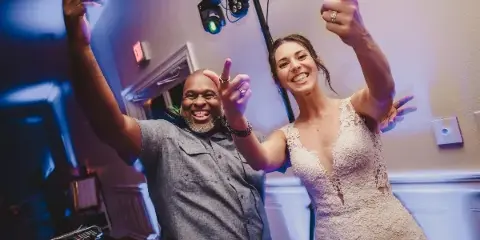 A smiling DJ and a bride in a white lace dress posing with raised hands in a venue with stage lighting and DJ equipment.