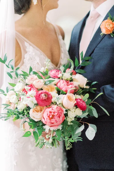 A close-up of a bride holding a large bouquet of pink, peach, and white flowers with greenery, standing next to a groom wearing a matching orange boutonniere.