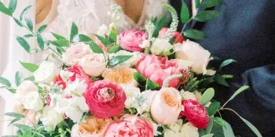 A close-up of a bride holding a large bouquet of pink, peach, and white flowers with greenery, standing next to a groom wearing a matching orange boutonniere.
