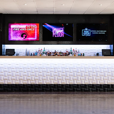 Interior view of a modern event venue featuring a long, white padded bar counter and digital screens displaying 'Welcome to the One and Only' and 'Dance Floor'.