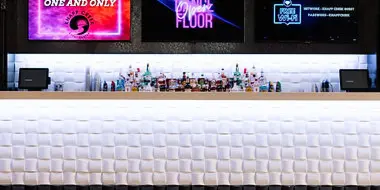 Interior view of a modern event venue featuring a long, white padded bar counter and digital screens displaying 'Welcome to the One and Only' and 'Dance Floor'.