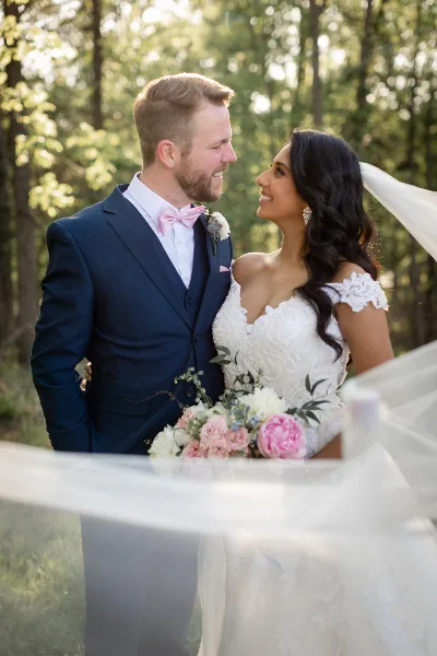 A bride and groom standing together outdoors in a wooded area, with the bride holding a bouquet of pink and white flowers.