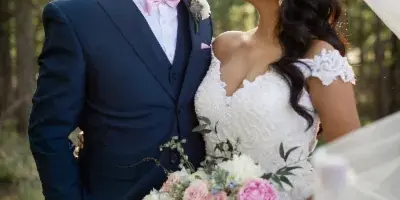 A bride and groom standing together outdoors in a wooded area, with the bride holding a bouquet of pink and white flowers.