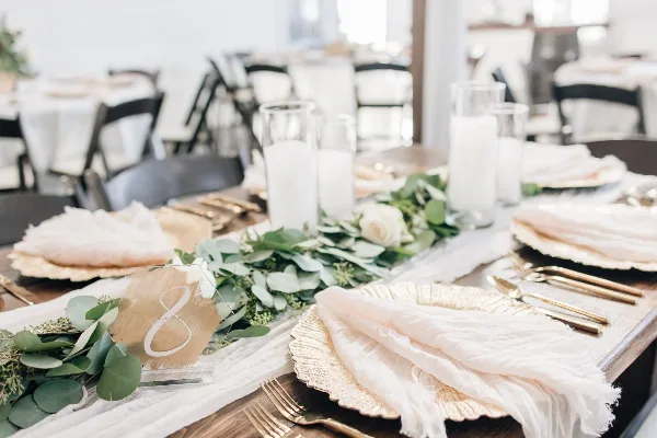 A close-up of a wedding table setting featuring gold plates, white napkins, and a greenery and white rose centerpiece.
