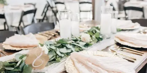 A close-up of a wedding table setting featuring gold plates, white napkins, and a greenery and white rose centerpiece.