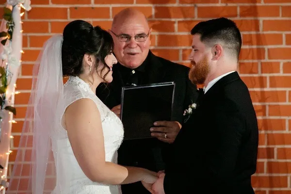 A wedding officiant in clerical attire stands between a bride and groom during a ceremony in front of a brick wall.