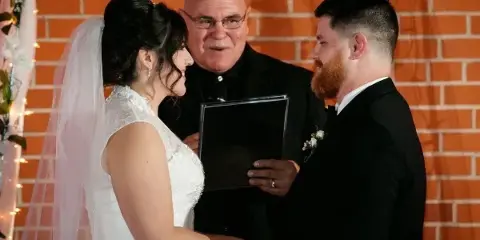 A wedding officiant in clerical attire stands between a bride and groom during a ceremony in front of a brick wall.