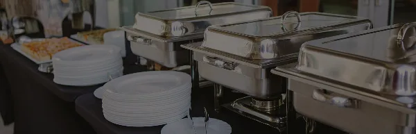 Catering buffet table with stainless steel chafing dishes, stacks of white plates, and prepared food items.