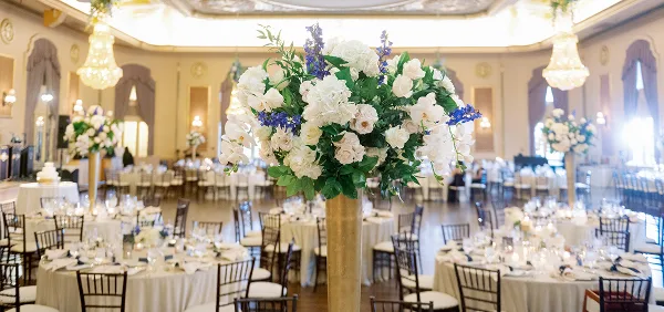 A grand ballroom decorated for a wedding with round tables, white linens, and a large floral centerpiece featuring white roses and blue delphiniums.