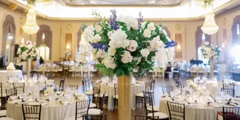 A grand ballroom decorated for a wedding with round tables, white linens, and a large floral centerpiece featuring white roses and blue delphiniums.