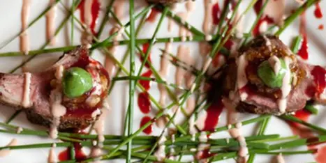 A close-up of a plated gourmet dish featuring meat, green garnish, and artistic sauce drizzles.