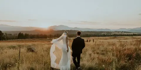 A bride and groom walking away from the camera through a golden grassy field with mountains in the background at sunset.