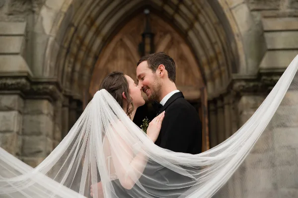 A bride and groom sharing a kiss in front of a stone archway, with the bride's long white veil flowing across the frame.