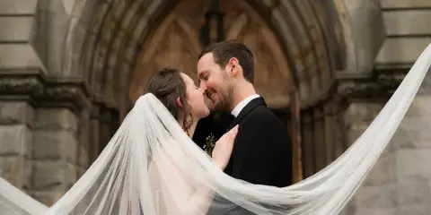 A bride and groom sharing a kiss in front of a stone archway, with the bride's long white veil flowing across the frame.