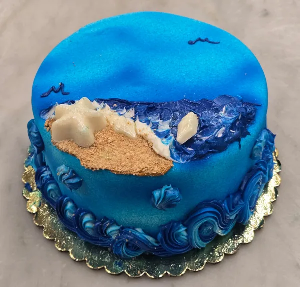 A round blue specialty cake with piped wave borders, a beach scene with sand and starfish, displayed on a gold cake board.