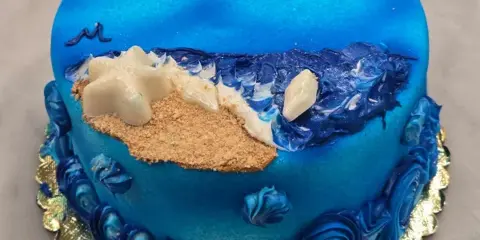 A round blue specialty cake with piped wave borders, a beach scene with sand and starfish, displayed on a gold cake board.