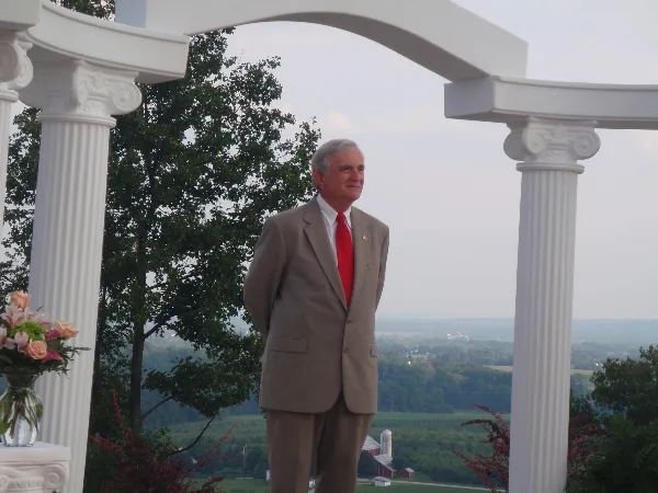 A man in a tan suit and red tie standing in a white wedding arch with flowers and a rural landscape in the background.