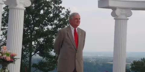 A man in a tan suit and red tie standing in a white wedding arch with flowers and a rural landscape in the background.