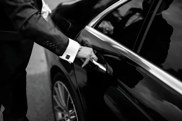 A professional chauffeur in a suit opening the door of a black luxury sedan.