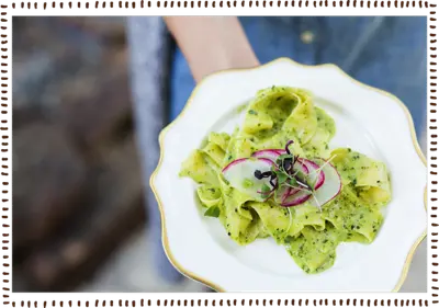 A plated appetizer with green pesto pasta, radish slices, and microgreens served on a white plate with a gold rim.