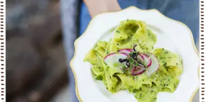 A plated appetizer with green pesto pasta, radish slices, and microgreens served on a white plate with a gold rim.