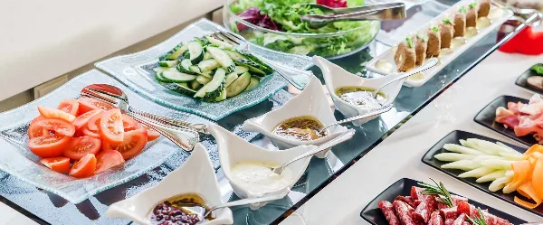 A catering buffet table featuring sliced tomatoes, cucumbers, a green salad, and various dips and appetizers.