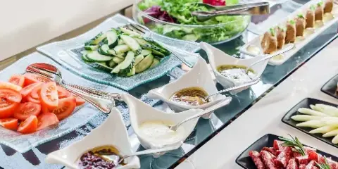 A catering buffet table featuring sliced tomatoes, cucumbers, a green salad, and various dips and appetizers.