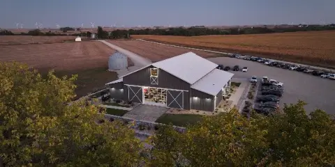 Aerial view of a modern barn-style wedding venue surrounded by rural farmland and a parking lot at dusk.