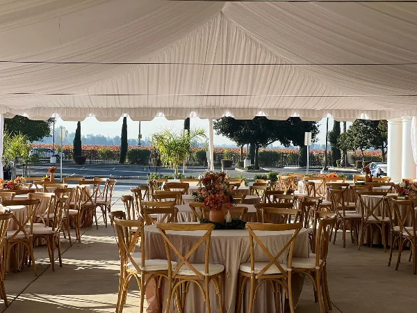 An outdoor event setup featuring round tables with beige tablecloths, wooden cross-back chairs, and floral centerpieces under a white draped tent.