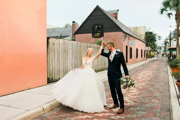 Bride in a white lace gown and groom in a dark suit dancing on a brick street while holding a bouquet.
