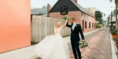 Bride in a white lace gown and groom in a dark suit dancing on a brick street while holding a bouquet.