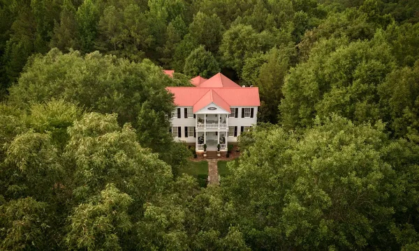 Aerial view of a white historic house with a red roof surrounded by dense green forest.