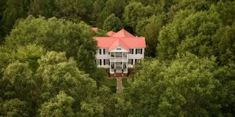 Aerial view of a white historic house with a red roof surrounded by dense green forest.
